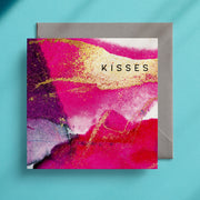 Square card with a pink and gold design and the caption "Kisses" in the top right corner on a grey envelope