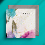 Square greeting card with pink, lilac and turquoise paint smears and gold splatters on a white background with the word "Hello" in the top right hand corner
