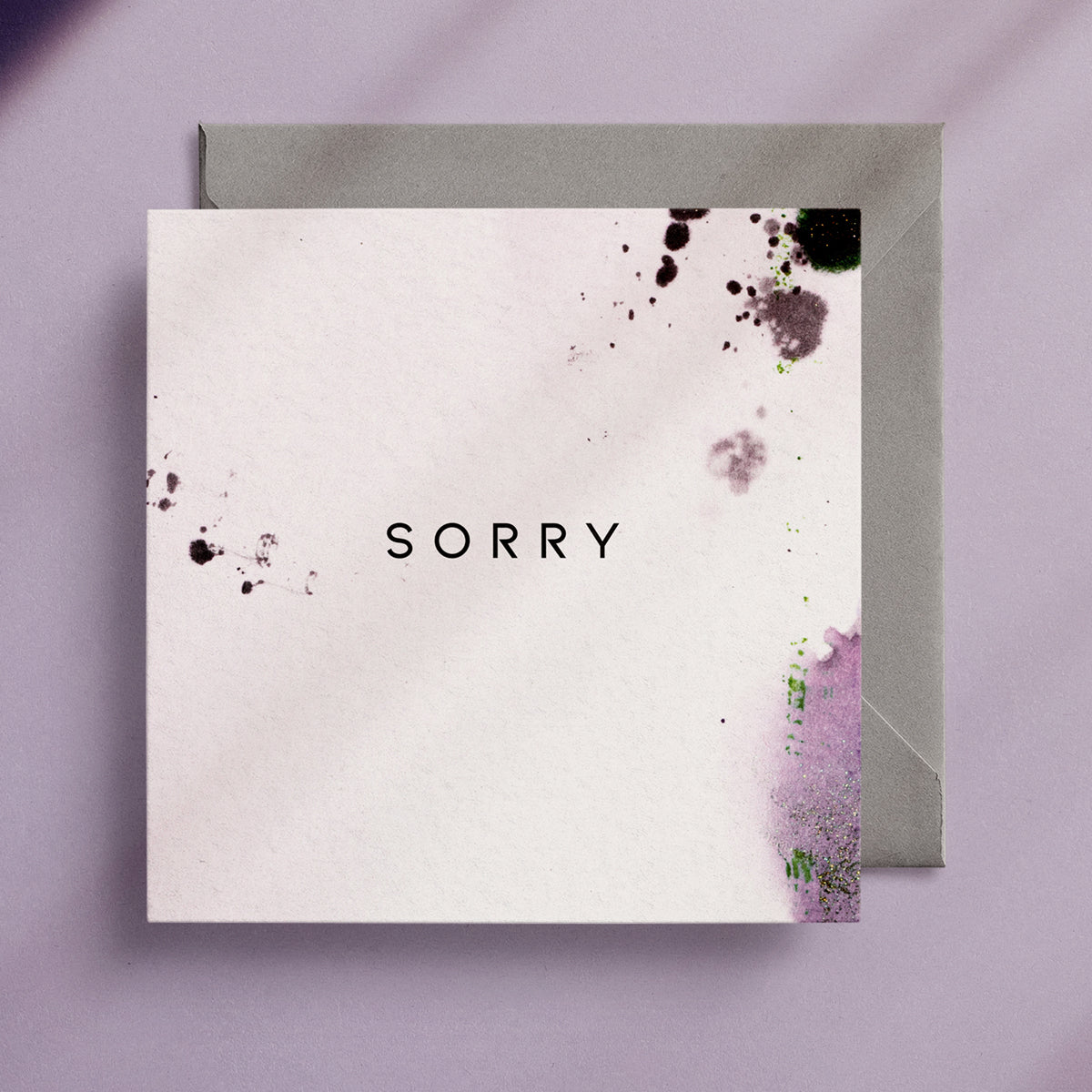 Square greeting card on top of a grey envelope with the word "Sorry" written in the middle