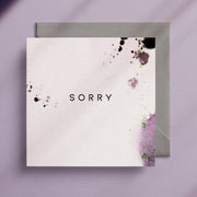 Square greeting card on top of a grey envelope with the word "Sorry" written in the middle